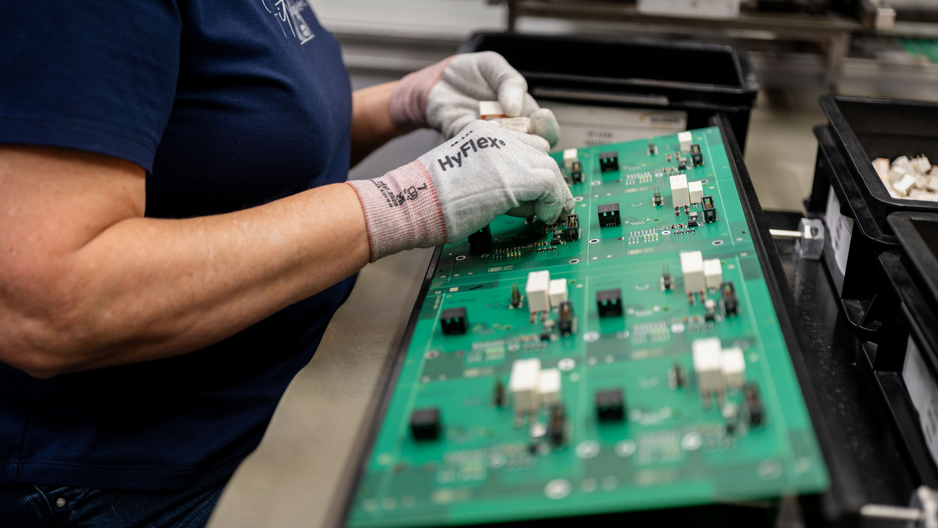 Leiterplatte beim Bestücken mit elektronischen Bauteilen bei der Geräte- und Systemmontage: Hände mit Spezialhandschuhen vor einem grünen Board mit weißen und schwarzen Bauteilen.
